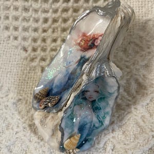 May include: Two decorative oyster shells featuring detailed illustrations of mermaids with flowing hair. The upper shell depicts a mermaid with reddish hair, while the lower shell shows one with teal hair. Both shells are accented with small seashells.