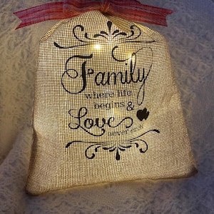 May include: A burlap bag with the words "Family where life begins & Love never ends" printed in black script. The bag is lit from within, and a red ribbon is tied at the top. Decorative item.