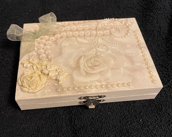 May include: A white wooden box with a hinged lid decorated with a pearl trim, a green ribbon, and a 3D floral design. The box has a silver latch.
