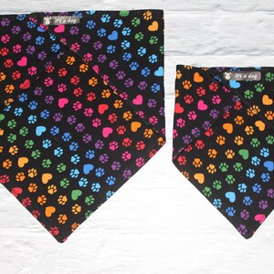 Könnte beinhalten: Zwei schwarze Hunde-Bandanas mit einem sich wiederholenden Muster aus bunten Pfotenabdrücken und Herzen. Die Bandanas haben ein Etikett mit der Aufschrift "it's a dog". Die Bandanas sind in verschiedenen Größen.
