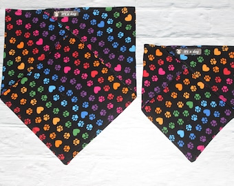 May include: Two black dog bandanas with a repeating pattern of colorful paw prints and hearts. The bandanas have a tag that reads "it's a dog". The bandanas are in different sizes.