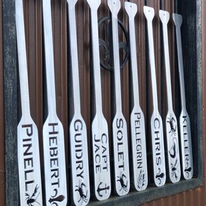 May include: Nine silver metal paddles with various names engraved on them, including "Pinell", "Hebert", "Guidry", "Capt Jace", "Solet", "Pellegrin", "Chris", and "Keller". Some paddles have a crab or anchor design.