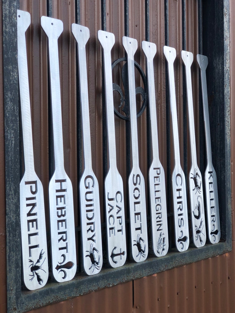 May include: Nine silver metal paddles with various names engraved on them, including "Pinell", "Hebert", "Guidry", "Capt Jace", "Solet", "Pellegrin", "Chris", and "Keller". Some paddles have a crab or anchor design.