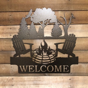 May include: Metal welcome sign featuring a silhouette of two chairs, a campfire, and trees. The sign reads "WELCOME".