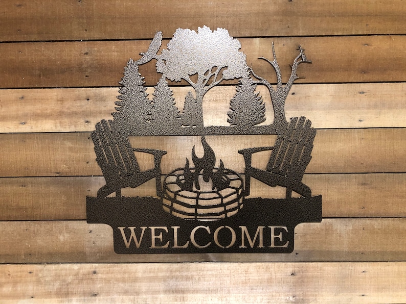 May include: Metal welcome sign featuring a silhouette of two chairs, a campfire, and trees. The sign reads "WELCOME".