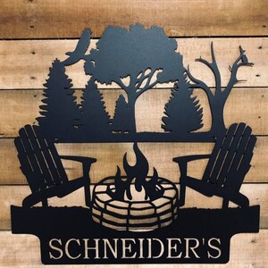 May include: Black metal silhouette of a campfire scene with two chairs, a fire pit, and trees. The text "SCHNEIDER'S" is below the scene.