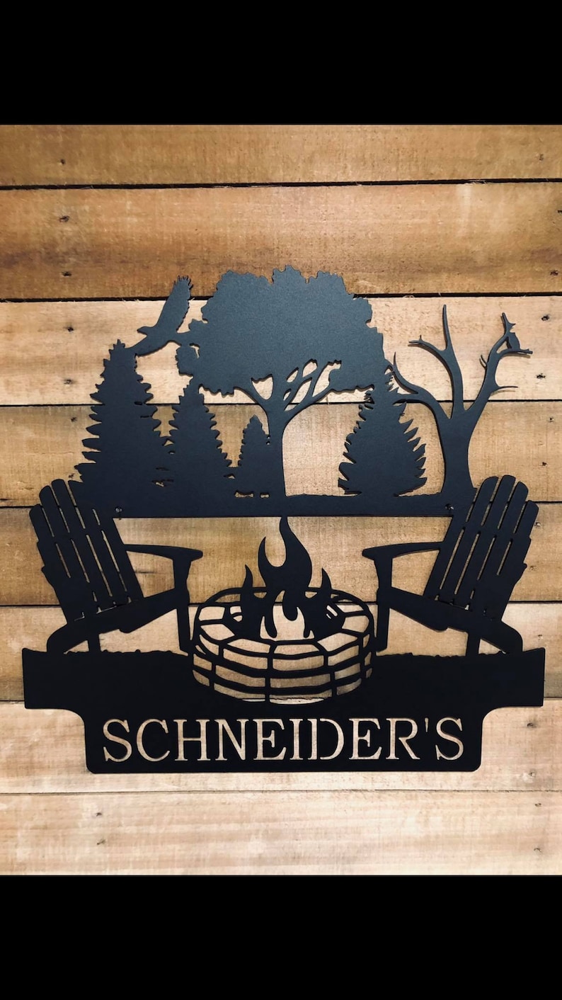 May include: Black metal silhouette of a campfire scene with two chairs, a fire pit, and trees. The text "SCHNEIDER'S" is below the scene.