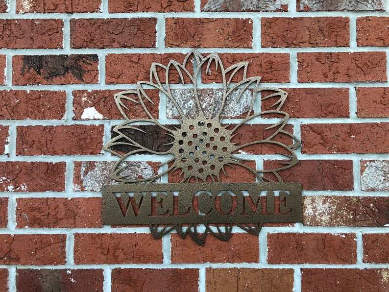 Op de afbeelding: Metalen zonnebloem welkomstbord met een bruine afwerking. Het bord is bevestigd aan een bakstenen muur.