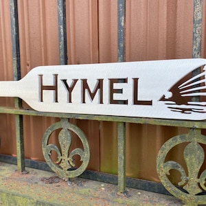 May include: White wooden paddle with the word "HYMEL" cut out and a silhouette of a fish with fins spread out.