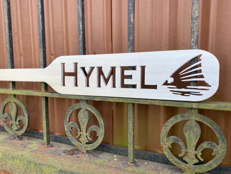 May include: White wooden paddle with the word "HYMEL" cut out and a silhouette of a fish with fins spread out.