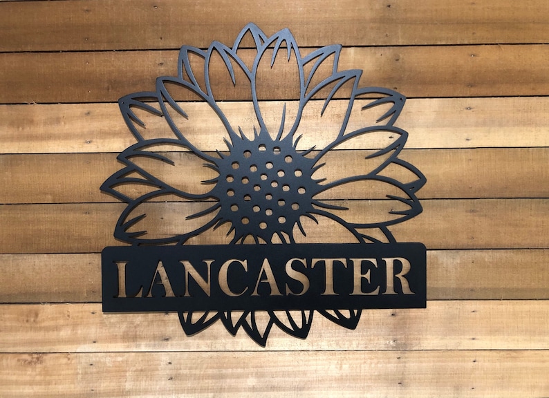 Op de afbeelding: Zwart metalen zonnebloemontwerp met de naam "LANCASTER" in gouden letters op een houten achtergrond.