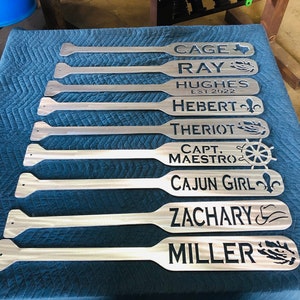 May include: Nine personalized wooden paddles with various names and designs. The names include Cage, Ray, Hughes Est 2022, Hebert, Theriot, Capt Maestro, Cajun Girl, Zachary, and Miller. Each paddle features a unique design, such as a Texas star, a fleur-de-lis, a ship's wheel, and a fishing hook.