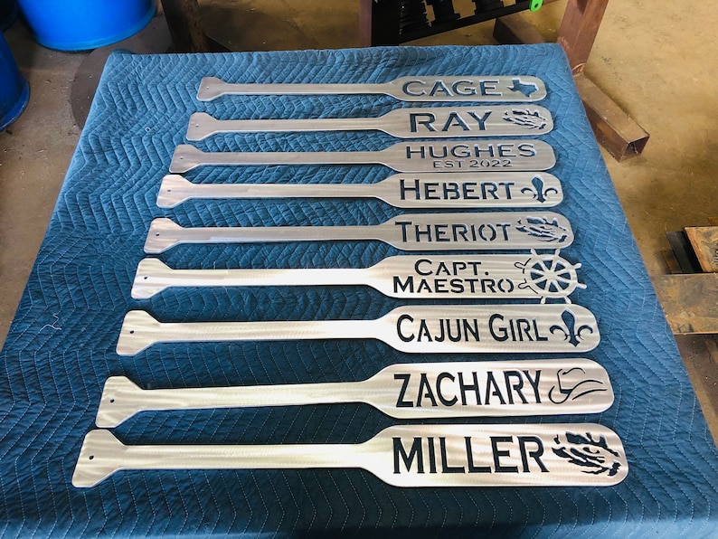 May include: Nine personalized wooden paddles with various names and designs. The names include Cage, Ray, Hughes Est 2022, Hebert, Theriot, Capt Maestro, Cajun Girl, Zachary, and Miller. Each paddle features a unique design, such as a Texas star, a fleur-de-lis, a ship's wheel, and a fishing hook.
