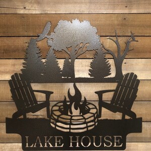 May include: Metal silhouette wall art featuring a campfire scene with two Adirondack chairs, a fire pit, and trees. The text "LAKE HOUSE" is displayed below the scene.