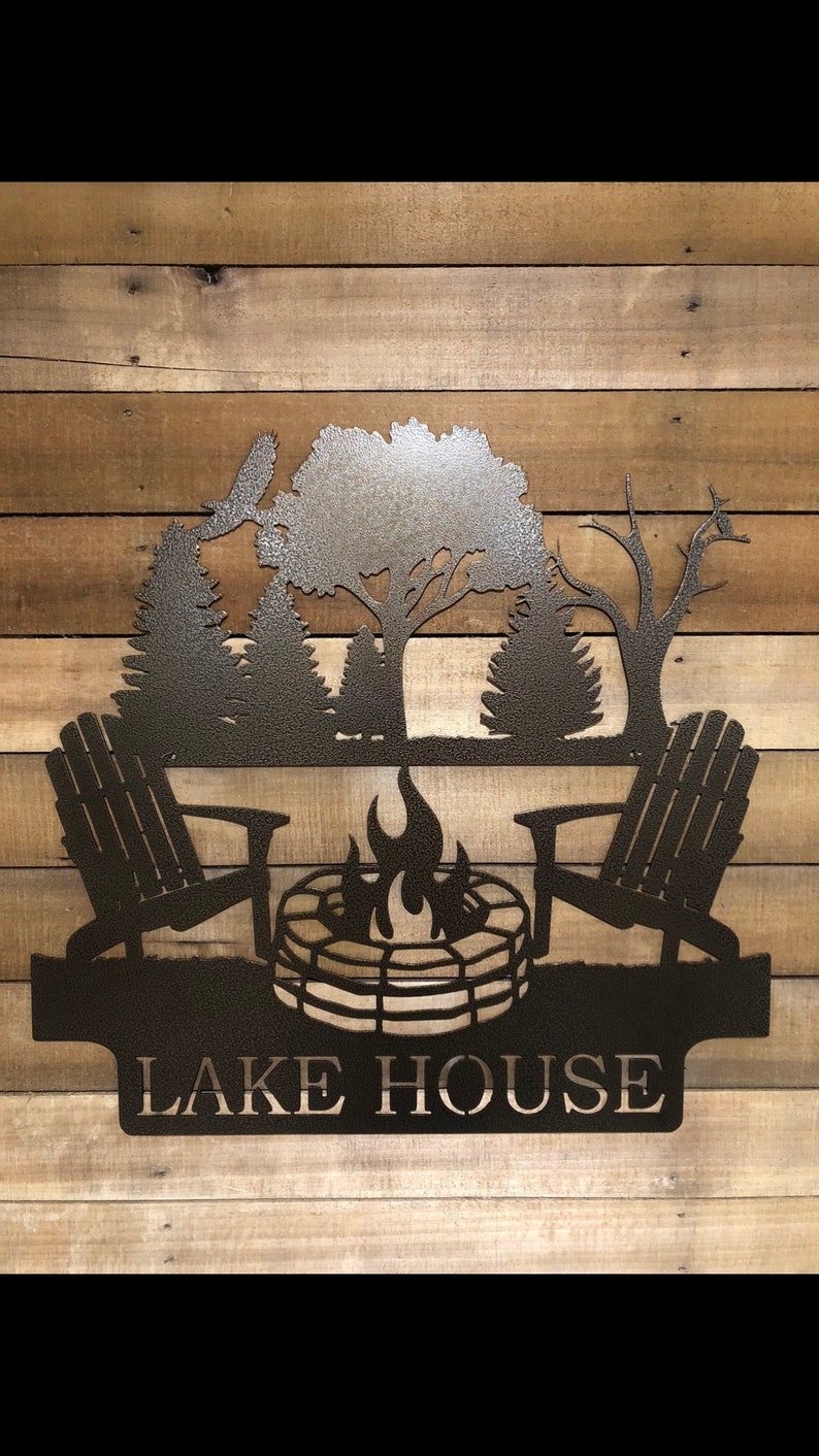 May include: Metal silhouette wall art featuring a campfire scene with two Adirondack chairs, a fire pit, and trees. The text "LAKE HOUSE" is displayed below the scene.