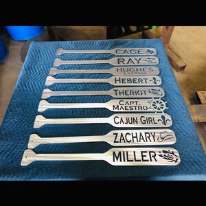 May include: A set of nine wooden paddles with personalised names and designs. The names include Cage, Ray, Hughes, Hebert, Theriot, Capt Maestro, Cajun Girl, Zachary, and Miller. Each paddle features a unique design, such as a Texas star, a fleur-de-lis, a ship's wheel, and a cowboy hat.