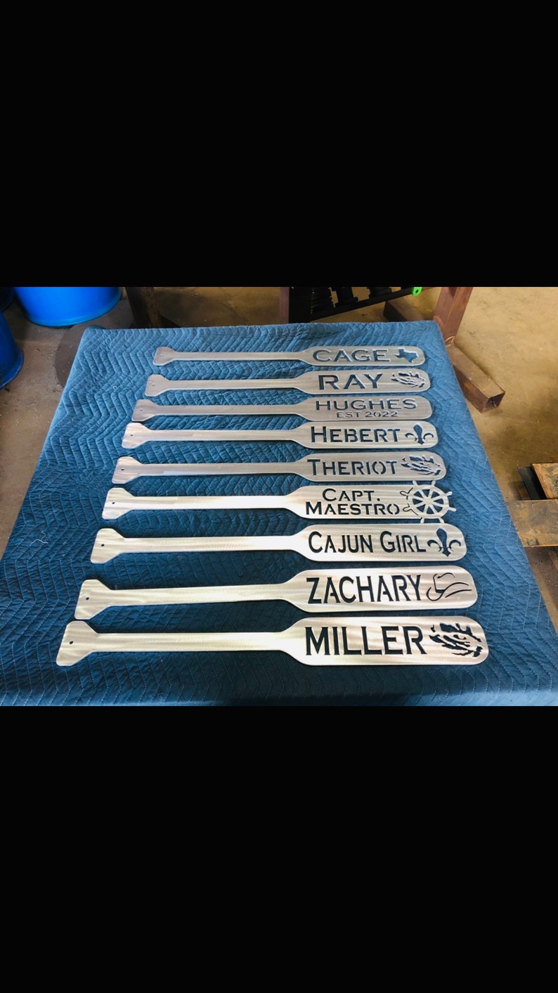 May include: A set of nine wooden paddles with personalised names and designs. The names include Cage, Ray, Hughes, Hebert, Theriot, Capt Maestro, Cajun Girl, Zachary, and Miller. Each paddle features a unique design, such as a Texas star, a fleur-de-lis, a ship's wheel, and a cowboy hat.
