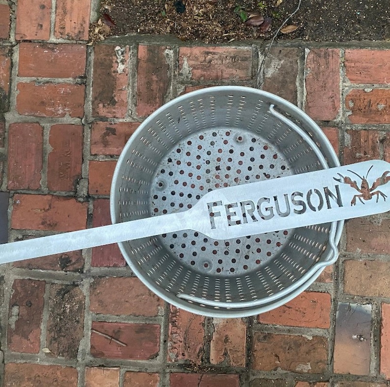 May include: A silver metal crawfish boil pot with a silver metal crawfish spatula with the name "FERGUSON" cut out. The pot and spatula are on a brick surface.