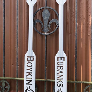 May include: Two white wooden paddles with the names "Boykin" and "Eubanks" cut out in the wood. Each paddle has a small square and compass symbol at the bottom.