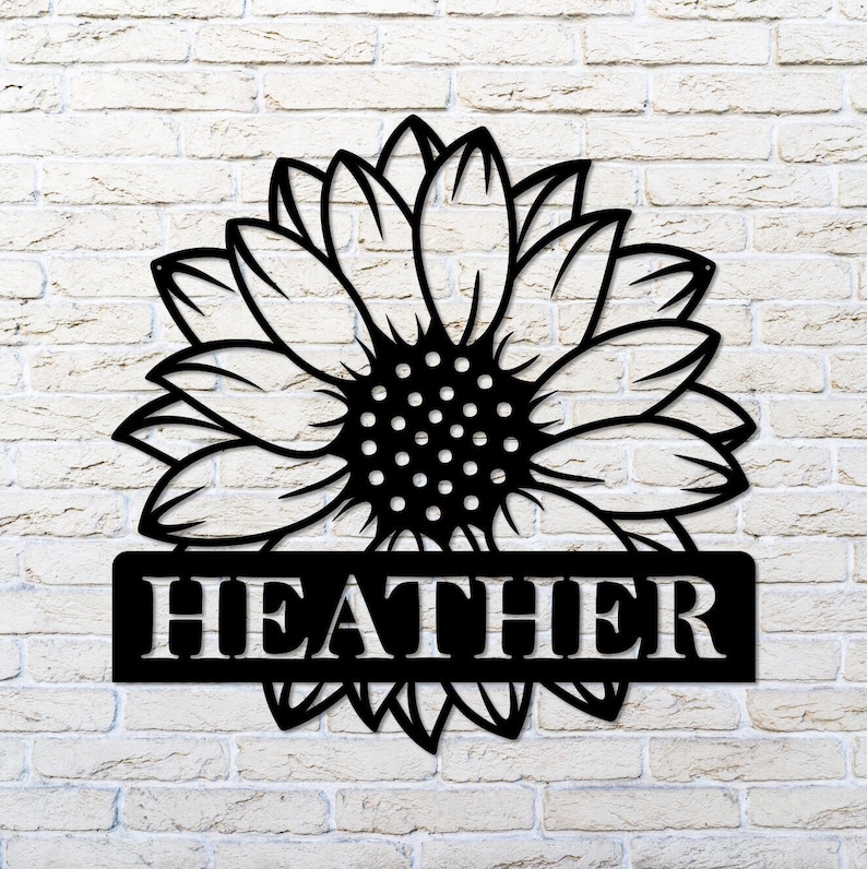 Op de afbeelding: Zwarte metalen zonnebloem met de naam "HEATHER" op een banner eronder. De zonnebloem is een populair ontwerp voor woonaccessoires en geschenken.