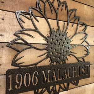Op de afbeelding: Een metalen zonnebloemontwerp met een zwarte afwerking. De zonnebloem heeft een lang, rechthoekig naambord met de tekst "1906 MALACHI ST" in wit.