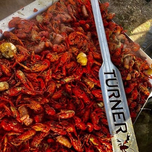 May include: A large metal serving utensil with the word "TURNER" engraved on it. The utensil is lying on top of a large tray of red crawfish.