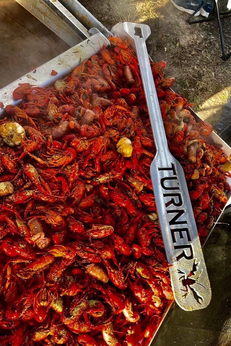 May include: A large metal serving utensil with the word "TURNER" engraved on it. The utensil is lying on top of a large tray of red crawfish.