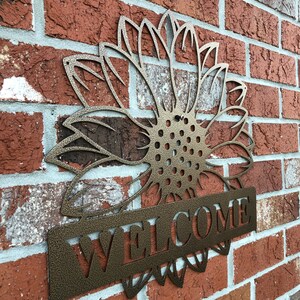 Op de afbeelding: Een metalen zonnebloem met een welkomstbord aan de onderkant. De zonnebloem is een uitsnijding en het bord is een massief stuk metaal. Beide zijn in brons geverfd.