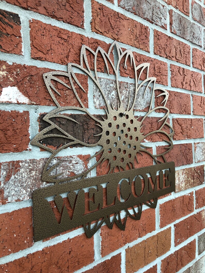 Op de afbeelding: Een metalen zonnebloem met een welkomstbord aan de onderkant. De zonnebloem is een uitsnijding en het bord is een massief stuk metaal. Beide zijn in brons geverfd.