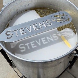 May include: A silver metal stirring utensil with the word "STEVENS" printed on it. The utensil is resting on top of a large pot filled with a white liquid.