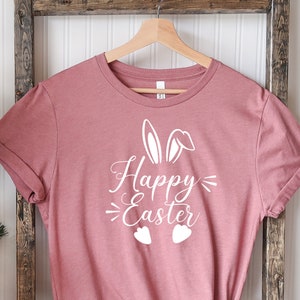 Op de afbeelding: Een roze T-shirt met een witte afbeelding van konijnenoren en de tekst "Happy Easter" op een houten ladder voor een kerstboom.