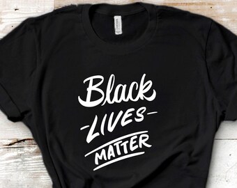 May include: Black t-shirt with white text that reads "Black Lives Matter" on a white wood background.