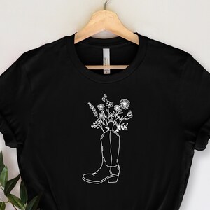 May include: Black t-shirt with a white outline of a cowboy boot filled with flowers. The design is centrally placed. The boot's details are clearly visible. The t-shirt is hanging on a wooden hanger.