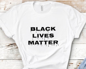 May include: White t-shirt with the text "BLACK LIVES MATTER" in black, with a dripping effect. The shirt is paired with light blue denim jeans. The items are arranged on a white wooden surface with small potted succulents.