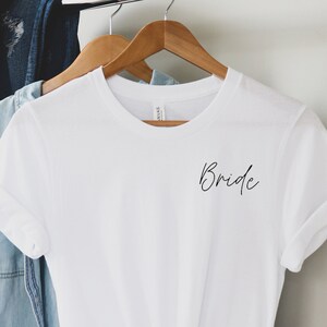 May include: White t-shirt with the word "Bride" written in black cursive on the front.