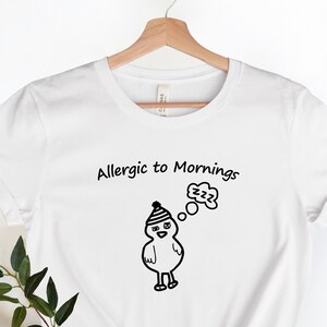 May include: White t-shirt with black text that reads "Allergic to Mornings". A cartoon chick wearing a hat is dreaming of sleeping, with a thought bubble above its head that says "zzz".