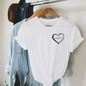 May include: White t-shirt with a black heart design and the word "ADOPT" inside the heart. The heart is outlined with paw prints.