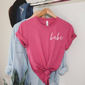 May include: A pink t-shirt with the word "babe" written in white cursive on the front.