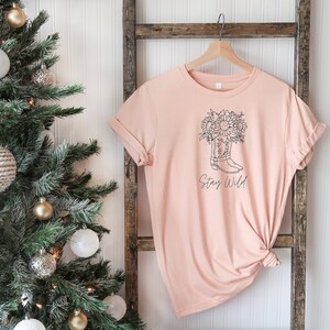 May include: A light peach-colored t-shirt with a black line drawing of a cowboy boot filled with sunflowers. The words "Stay Wild" are written below the boot. The shirt is hanging on a wooden hanger.