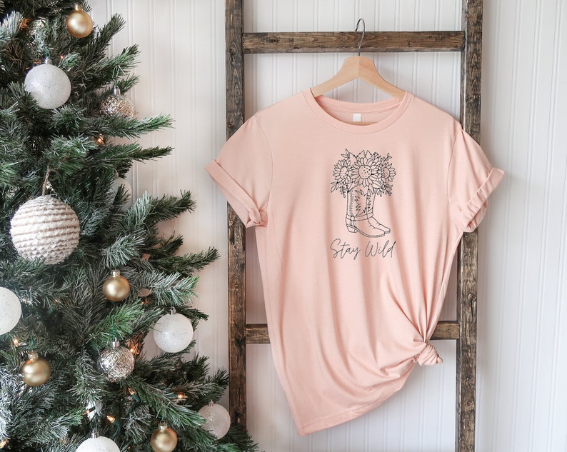 May include: A light peach-colored t-shirt with a black line drawing of a cowboy boot filled with sunflowers. The words "Stay Wild" are written below the boot. The shirt is hanging on a wooden hanger.