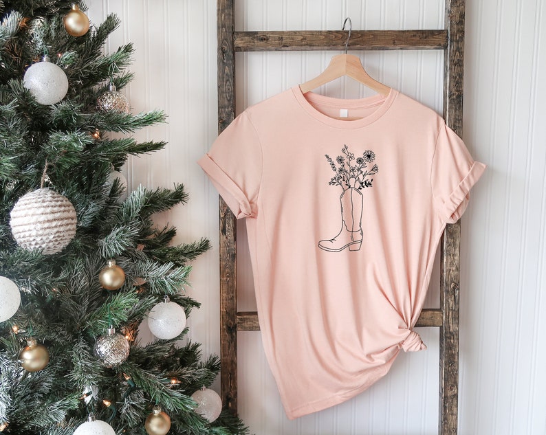 May include: A pale peach t-shirt with a black outline of a cowboy boot filled with flowers. The tee is hanging on a wooden ladder, with a Christmas tree in the background.