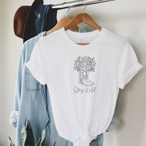 May include: White t-shirt with a black and white graphic of a cowboy boot filled with sunflowers. The text "Stay Wild" is below the boot. The shirt is hanging on a wooden hanger with other clothing items.
