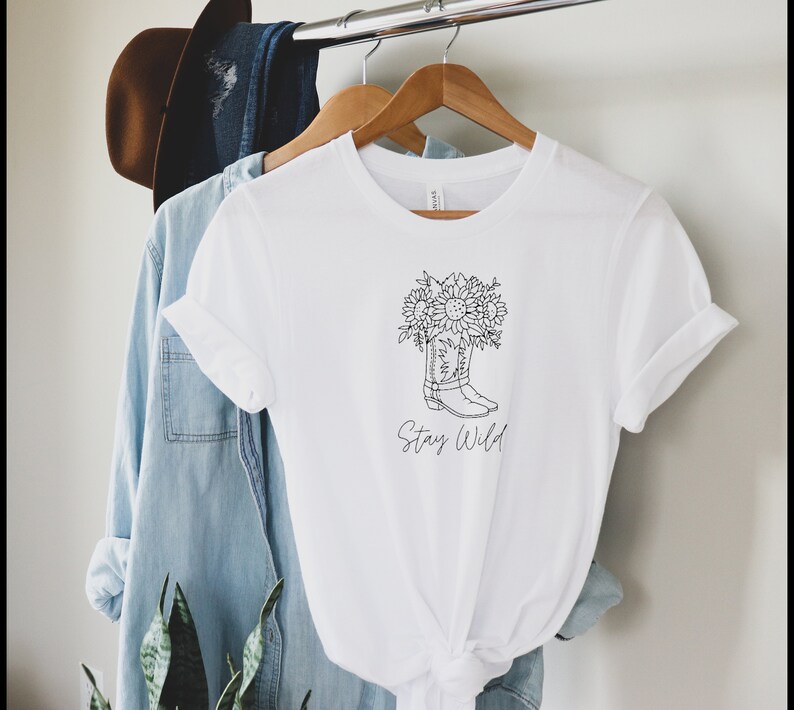May include: White t-shirt with a black and white graphic of a cowboy boot filled with sunflowers. The text "Stay Wild" is below the boot. The shirt is hanging on a wooden hanger with other clothing items.