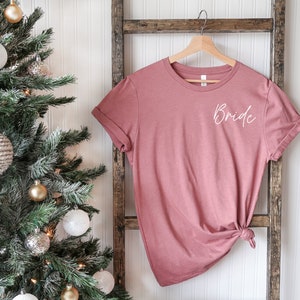 May include: A dusty rose colored t-shirt with the word "Bride" written in white script on the front. The t-shirt is hanging on a wooden ladder.
