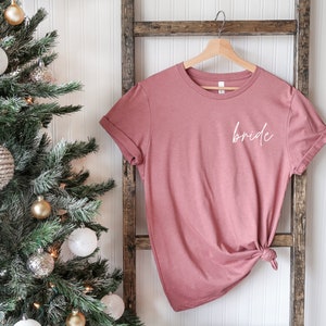 May include: A dusty rose colored t-shirt with the word "bride" written in white script on the front. The shirt is hanging on a wooden ladder against a white wall.