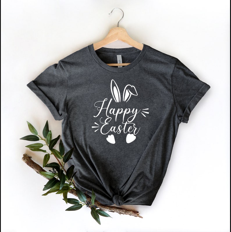 Op de afbeelding: Een donkergrijs T-shirt met een wit konijnenorenontwerp en de tekst "Happy Easter" gedrukt op de voorkant.