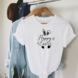 Op de afbeelding: Wit T-shirt met een zwart grafisch ontwerp met de tekst "Happy Easter" met konijnenoren en harten.