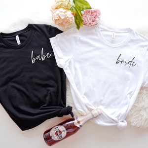 Op de afbeelding: Twee T-shirts, een zwart en een wit, met de woorden "babe" en "bride" in wit schrift op de voorkant.