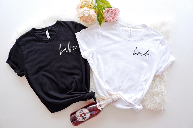 Op de afbeelding: Twee T-shirts, een zwart en een wit, met de woorden "babe" en "bride" in wit schrift op de voorkant.