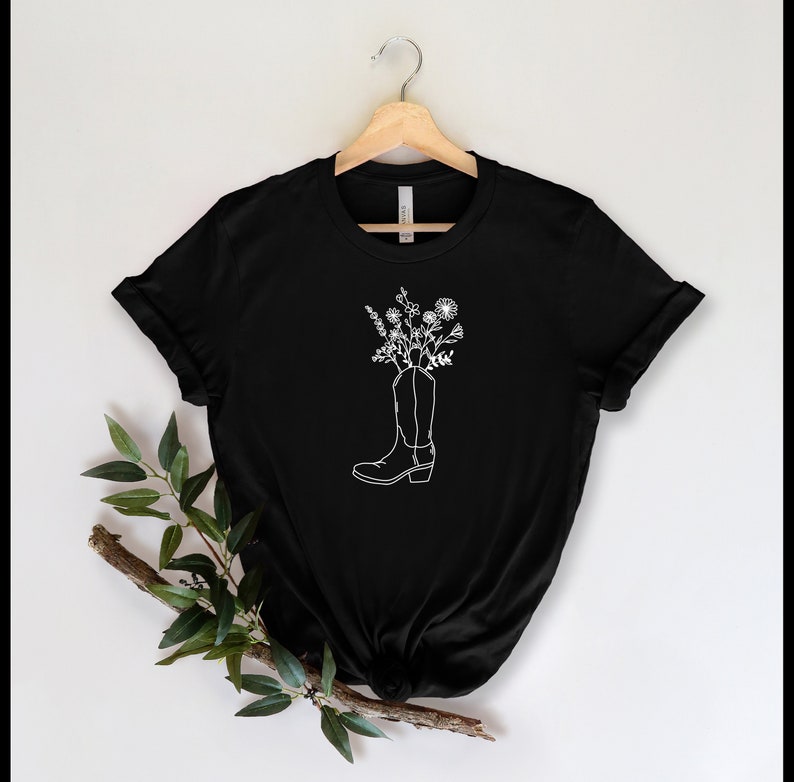 May include: Black t-shirt with a white outline of a cowboy boot filled with flowers. The design is centrally placed. The boot's details are clearly visible. The t-shirt is hanging on a wooden hanger.